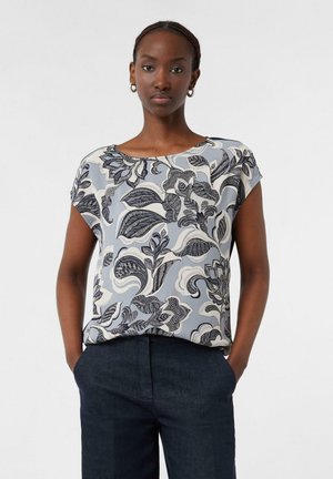 Kobieta ubrana w bluzkę z krótkim rękawem z dużym czarno-białym kwiatowym wzorem oraz ciemne spodnie z wysokim stanem, ręce w kieszeniach, neutralne tło.