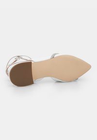 Chaussure à talon haut blanche avec un orteil pointu, des brides de cheville transparentes et une semelle en caoutchouc texturée. Comprend un détail de sangle décoratif en argent.