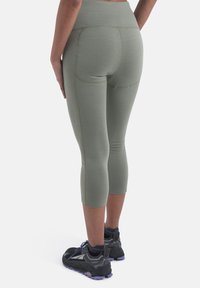 Kvinde iført olivengrønne cropped leggings og sorte trail running sko med blå såler, stående med hænderne hvilende på lårene.