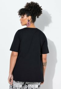 Černé oversized tričko s krátkým rukávem, kulatým výstřihem a hladkým bavlněným povrchem; modelka má na sobě vzorované kalhoty a velké náušnice.
