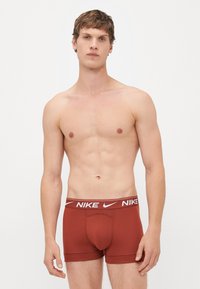 Bruine katoenen boxershorts met een witte Nike-logo tailleband, met een strakke pasvorm en een gladde textuur. Model staat rechtop, shirthless.