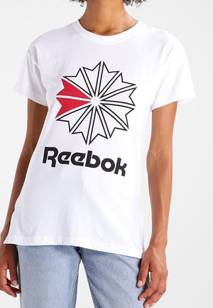 Osoba ubrana w białą koszulkę Reebok z czarno-czerwonym geometrycznym logo gwiazdy, zestawioną z jasnoniebieskimi dżinsami.