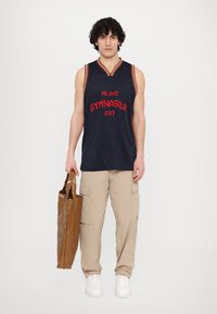 Námořnický síťovaný basketbalový dres s červeným grafickým textem, béžové cargo kalhoty a lesklá hnědá taška. Model má na sobě bílé boty.