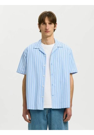 Homme portant une chemise à manches courtes à rayures verticales bleu clair et blanc par-dessus un t-shirt blanc avec un jean bleu, debout devant un fond uni.