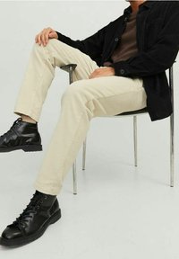 Pantaloni in corduroy crema con una texture a coste, abbinati a una camicia nera con bottoni e stivali alla caviglia neri con lacci, seduto su una sedia d'argento.