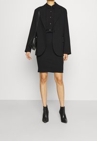 Blazer negro a medida sobre una camisa negra de botones y una falda negra ajustada, combinado con botas negras hasta el tobillo y un pequeño bolso cruzado negro.