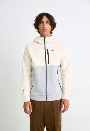 Haut à capuche zippé en crème clair et gris avec des accents noirs, à manches longues et poches latérales, fabriqué dans un tissu lisse et extensible.