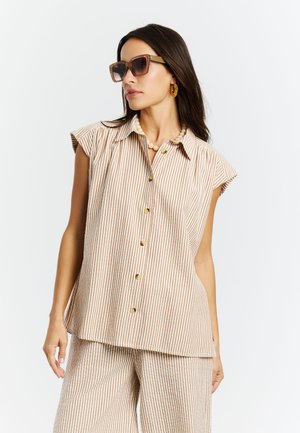Femme portant une chemise sans manches beige et blanche à rayures boutonnée et un pantalon large assorti, accessoirisée avec des lunettes de soleil, des boucles d'oreilles créoles et un collier de perles.