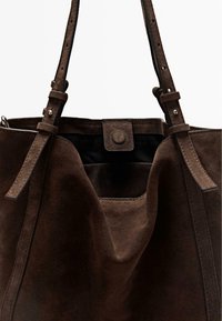 Donkerbruine suède boodschappentas met verstelbare schouderbanden en een magnetische drukknopsluiting, gedeeltelijk open weergegeven tegen een witte achtergrond.