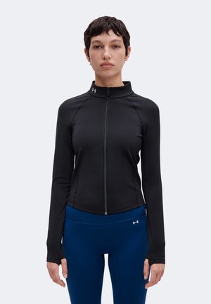 Personne aux cheveux courts et foncés portant une veste noire ajustée zippée et des leggings de sport bleus avec des logos de marque sur le col et la taille.