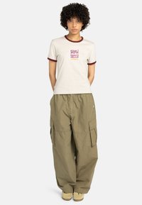 Crème t-shirt met bordeauxrode trim en grafische tekst, gecombineerd met losvallende olijfgroene cargobroek en beige schoeisel.