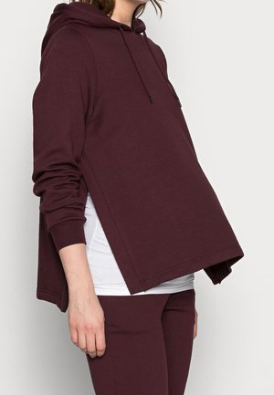 Capuchon sweatshirt in diep bordeaux, met een ontspannen pasvorm, lange mouwen en een gesplitste onderrand. Gemaakt van zachte, gestructureerde stof die over een wit shirt valt.