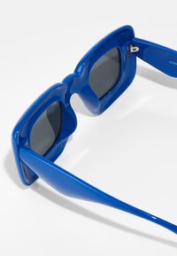 Ochelari de soare albaștri cu un cadru din plastic lucios, de formă rectangulară supradimensionată și lentile întunecate. Prezintă un design ondulat unic la sprâncene.
