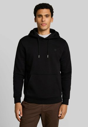 Junger Mann mit lockigem Haar, der einen schwarzen Kapuzenpullover und braune Hose trägt, steht vor einem schlichten hellgrauen Hintergrund.