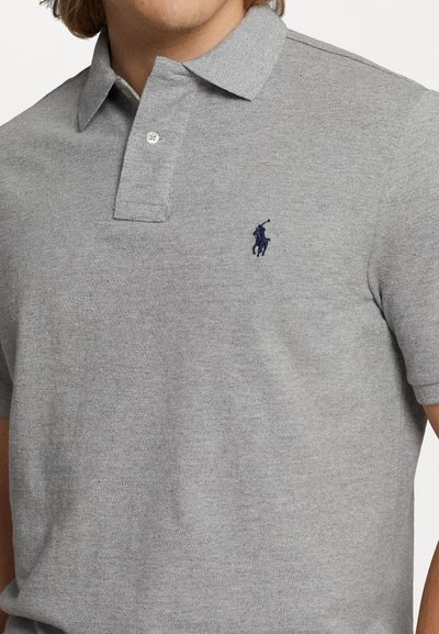 Pilkos spalvos polo marškiniai, pagaminti iš tekstūrinio medvilnės, su trimis sagomis priekyje ir tamsiai mėlynu žirgo logotipu ant kairės krūtinės. Trumpomis rankovėmis.