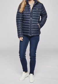 Granatowa kurtka puchowa z poziomym pikowaniem, pełnym zamkiem błyskawicznym i stójką. Połączona z ciemnoniebieskimi dżinsami i białymi trampkami.