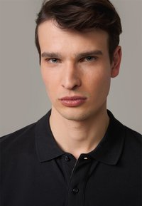 Schwarzes Polohemd mit klassischem Kragen und zwei Knöpfen, das ein klares Design, eine strukturierte Stoffqualität und minimale Details aufweist.