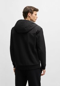 Felpa nera con una sezione superiore impermeabile, dotata di cappuccio regolabile con cordini, polsini a coste e una finitura liscia e opaca.