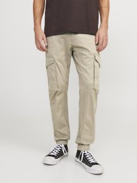 Pantaloni cargo bej cu manșete elastice, multiple buzunare laterale și material din bumbac. Asortați cu adidași negri cu sireți albi.