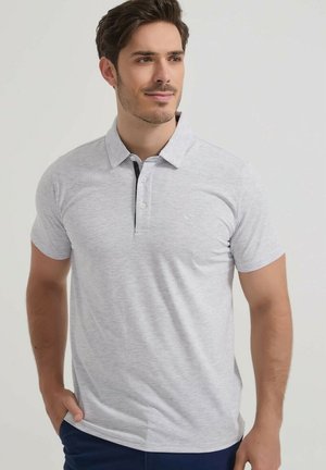 Homme portant un polo gris clair à manches courtes avec des boutons, debout avec une main dans la poche, regardant sur le côté.