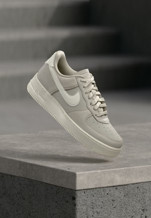 Μπεζ αθλητικό παπούτσι Nike Air Force 1 που αιωρείται πάνω από μια τσιμεντένια ακμή με θολές γκρίζες σκάλες στο φόντο.