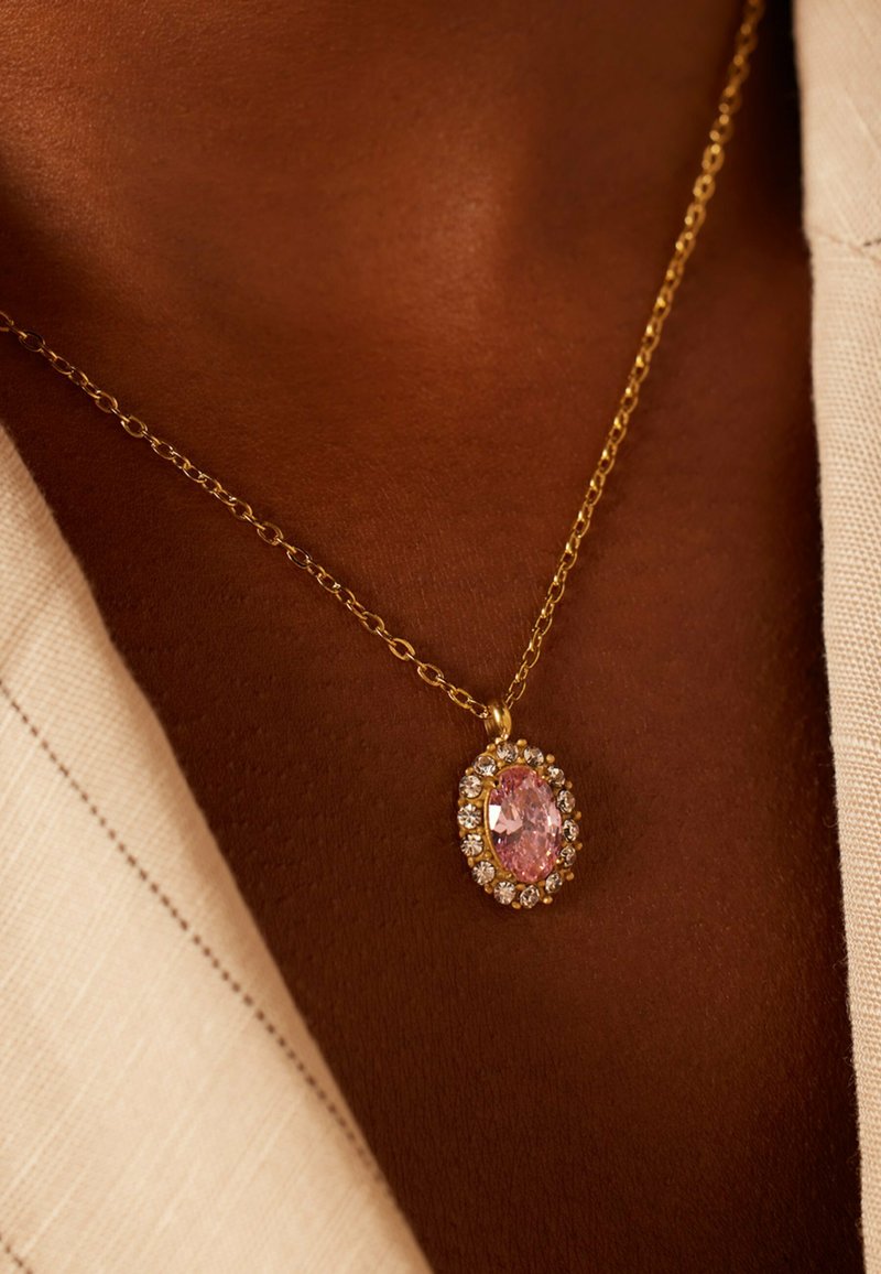 Gouden ketting met een ovale roze edelsteen omringd door kleine heldere stenen, rustend tegen een bruine huidkleur.
