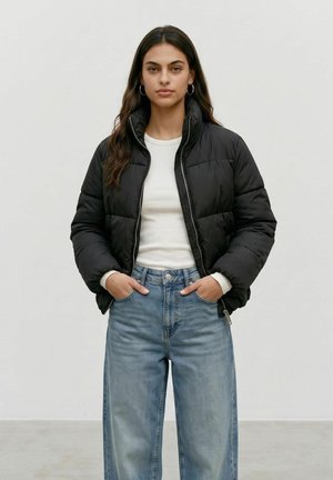 Frau mit langen dunklen Haaren, die eine schwarze Daunenjacke, ein weißes Hemd und hellblaue Jeans trägt, steht mit den Händen in den Taschen.
