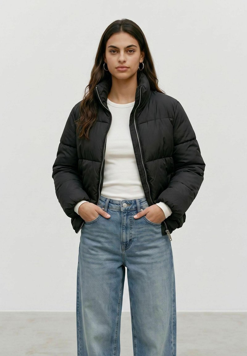 Femme aux longs cheveux foncés portant une doudoune noire, une chemise blanche et un jean bleu clair, debout les mains dans les poches.