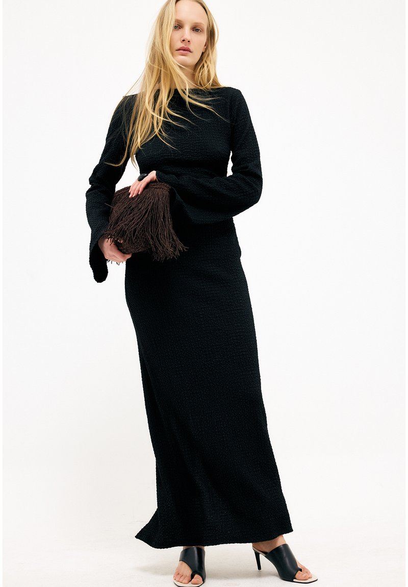 Nainen, joka pukeutuu pitkään mustaan teksturoituun mekkoon, jossa on levenevät hihat. Hän pitää kädessään ruskeaa tupsullista clutch-laukkua ja jalassaan mustat avokkaat, joissa on korko ja avoin kärki.