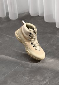 Stivale alto beige con tessuto testurizzato e pelle liscia, dotato di lacci neri, punta rinforzata e suola robusta per una maggiore aderenza.