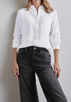 Persona con una camisa blanca texturizada de botones y mangas remangadas, metida dentro de unos vaqueros negros de cintura alta, de pie frente a una pared clara.