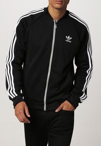 Black zip-up track jacket with white three-stripe design on sleeves. Features smooth fabric and an embroidered logo on the chest.