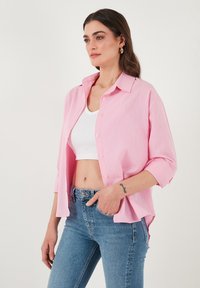 Light pink button-up shirt with rolled sleeves, worn over a white crop top. Model paired with light blue high-waisted jeans.