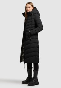 Cappotto lungo imbottito nero con motivo a righe orizzontali, chiusura con zip e cappuccio regolabile. Indossato sopra pantaloni neri e stivali chunky con lacci.