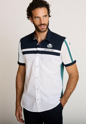 Homme aux cheveux bouclés foncés portant une chemise blanche et bleu marine à manches courtes avec le logo d'une équipe de course, les mains relâchées le long du corps.