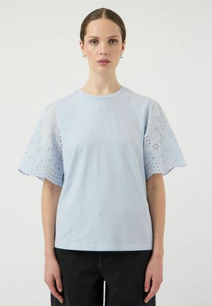 Vrouw met een lichtblauw shirt met oogjeskant gedetailleerde korte mouwen, gecombineerd met een zwarte broek, staand tegen een effen witte achtergrond.