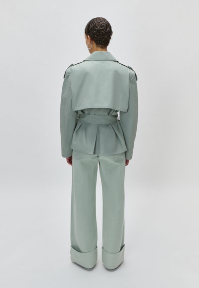 Άτομο με κοντά σγουρά μαλλιά που φοράει ανοιχτό πράσινο σακάκι με ζώνη και παντελόνι με φαρδιά πόδια, φαίνεται από πίσω μπροστά σε απλό φόντο.
