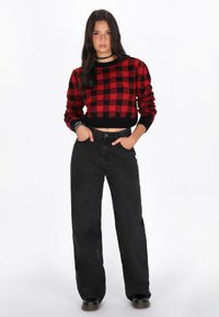 Rot-schwarz kariertes, gestricktes, kurzes Sweatshirt mit langen Ärmeln, kombiniert mit hochtaillierten schwarzen weit geschnittenen Hosen. Schwarze Stiefel.