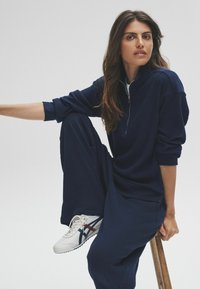 Femme assise sur un tabouret en bois, portant un sweat zippé en côtelé marine, un pantalon assorti et des baskets blanches avec des rayures marines et rouges.