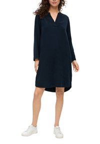 Granatowa sukienka z dekoltem w serek, długimi rękawami i luźnym krojem. Miękka tkanina o gładkiej fakturze, sięgająca do połowy uda. Noszona z białymi trampkami.