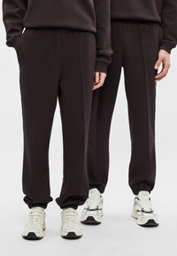 Tmavohnedé tepláky s voľným strihom, elastickým pásom a manžetami. Hladká látka s záhybmi; v kombinácii so svetlými teniskami.