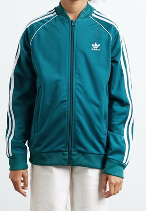 Person trägt eine petrolfarbene Adidas-Zip-up-Jacke mit weißen Streifen an den Ärmeln und Logo auf der Brust, kombiniert mit weißen Hosen.