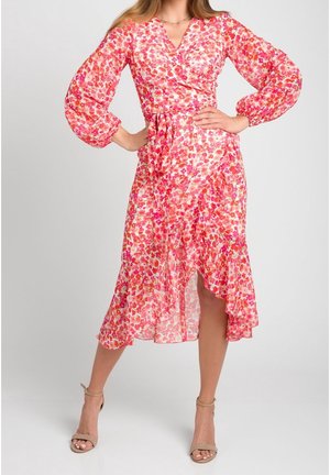 Mujer con un vestido cruzado floral en tonos rosa y rojo con mangas largas abullonadas y tacones beige con correa en el tobillo, de pie con las manos en las caderas.