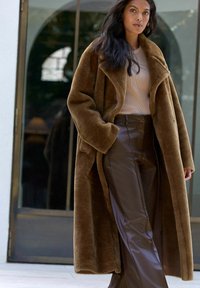 Brun faux fur frakke over en lys beige top og brune læderbukser; har en bred krave og forlommer. Glat tekstur hele vejen igennem.