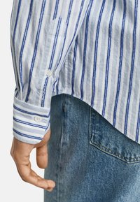 Blau-weiß gestreiftes Langarmhemd mit Knopfmanschetten, aus leichtem Stoff, kombiniert mit blauen Jeans mit einer Gesäßtasche.