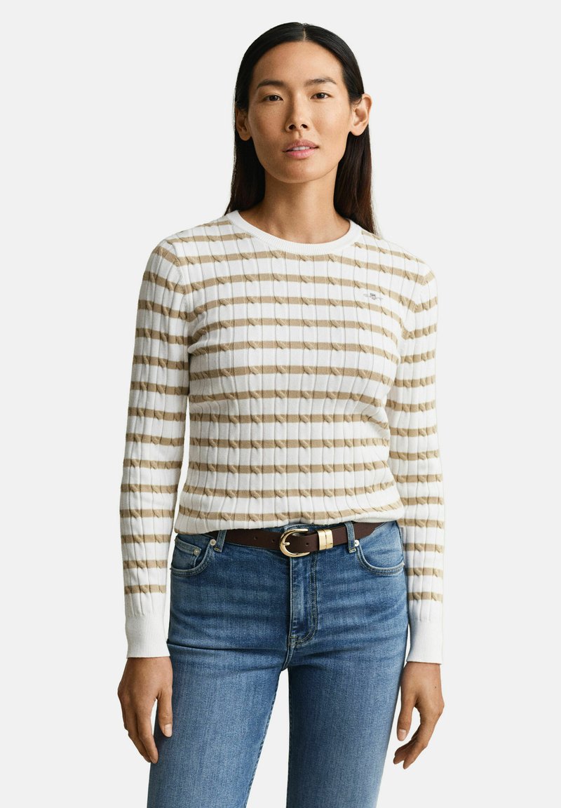 Kvinde iført hvid og beige stribet kabelstrikket sweater, blå jeans og brunt bælte, vendt mod kameraet med neutralt udtryk.