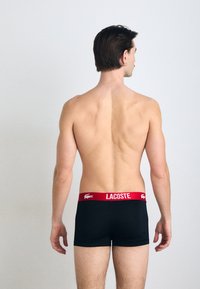 Crne bokserice s crvenim pojasom s Lacoste logotipom, koje se odlikuju uskim oblikom i glatkom teksturom, prikazane s leđa.
