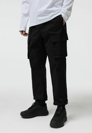 Osoba ubrana w czarne spodnie typu cargo z bocznymi kieszeniami, czarne trampki i białą koszulę z długim rękawem, stojąca na jednolitym jasnym tle.