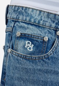 Blue denim jeans feature a back pocket with white embroidery, metal buttons, and contrasting orange stitching. Smooth, textured fabric visible.