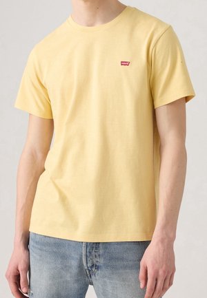Hombre con camiseta de manga corta Levi's de color amarillo claro y vaqueros azul claro, de pie frente a un fondo neutro.